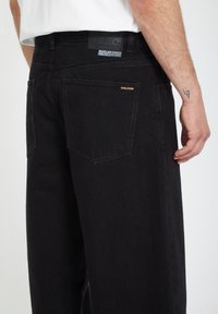 Black denim pants with a loose fit, featuring a prominent back pocket and a subtle logo in gold on one pocket. Textured fabric.