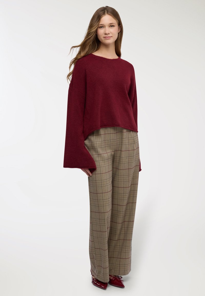 Maglione bordeaux lavorato a maglia con maniche larghe, abbinato a pantaloni beige a quadri con accenti rossi e una vestibilità comoda. Scarpe rosse lucide completano il look.