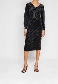 Svart klänning med lång ärm och V-ringning, tillverkad av glänsande tyg med ett prickigt mönster. Slim fit kjol som slutar strax under knäet, i kombination med svarta sandaletter med remmar.