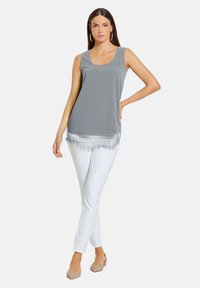 Grauer ärmelloser Oberteil mit Rundhalsausschnitt, versehen mit einem dekorativen Spitzenbesatz am Saum und kombiniert mit weißen Hosen. Glatte Stoffoberfläche.