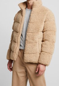 Beige pufferjack met een getextureerde, harige buitenkant, ritsluiting, hoge kraag en gestikte horizontale panelen. Draagt over een grijs T-shirt.