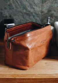 Brown leather toiletry bag with a zipper closure, featuring a smooth texture and rounded, structured shape. Minimalist design, no visible patterns.