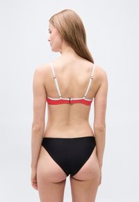 Γυναίκα που στέκεται με την πλάτη γυρισμένη, φορώντας μπικίνι με κόκκινο και λευκό τοπ και μαύρο κάτω μέρος, μπροστά σε μονόχρωμο φόντο.