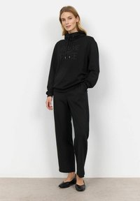 Felpa nera con la scritta "FEMME", cappuccio con cordini, abbinata a pantaloni neri larghi e scarpe nere piatte, su uno sfondo neutro.