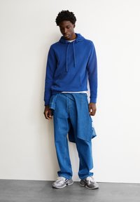 Blauer Kapuzenpullover mit einer Fronttasche, kombiniert mit locker sitzenden Jeans mit einem Einsatz. Das Modell trägt graue Turnschuhe mit weißen Schnürsenkeln.