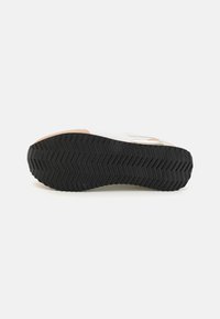 Hvit sneaker med en teksturert svart gummisåle, som har et sikksakkmønster, og en lys brun detalj på helen.