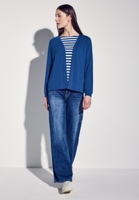 Blå öppen cardigan över en vit och marinrandig topp, kombinerad med vida blå jeans och vita sneakers. Slät tygkvalitet och löst passform.