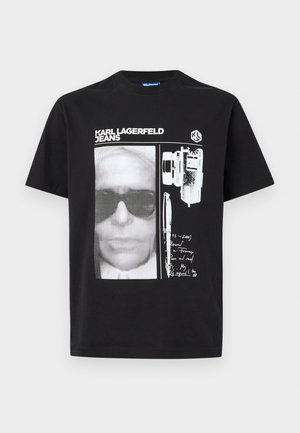 Melna T-krekls ar baltu pustoņu grafiku, kurā attēlota seja ar saulesbrillēm, kamera, pildspalva, roku rakstīts teksts un "KARL LAGERFELD JEANS" logotips.
