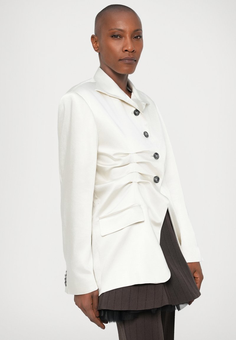 Vit skräddarsydd kavaj med framträdande knappar, rynkade detaljer framtill och en sidficka, i kombination med en plisserad mörk kjol.