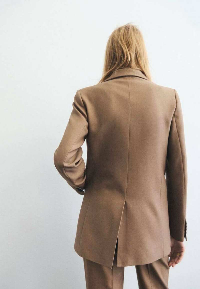 Personne aux longs cheveux blonds portant une veste de costume marron sur mesure et un pantalon assorti, debout devant un fond clair uni, vue de dos.