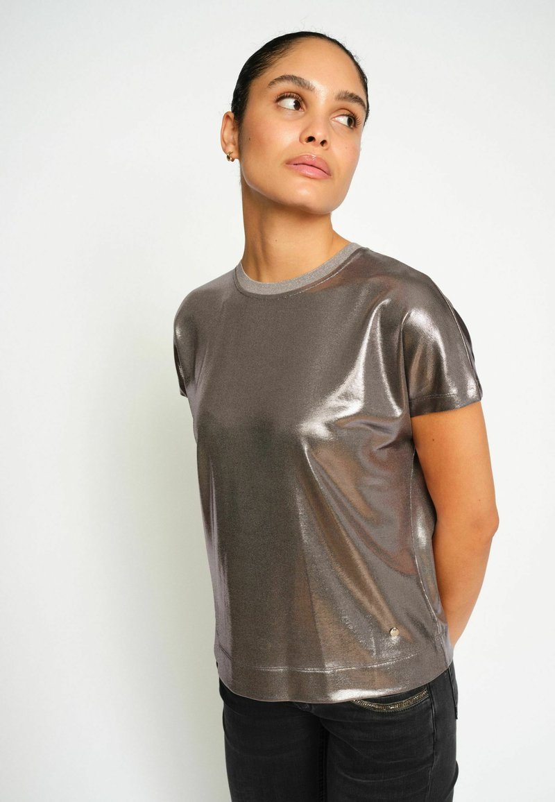 Tricou scurt, de culoare gri metalic, cu guler rotund, textura netedă și o strălucire subtilă, asortat cu blugi negri. Are un mic logo la margine.