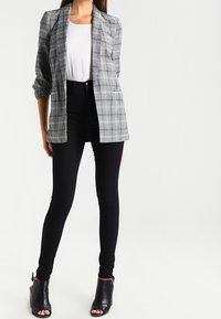Šedý kostkovaný blejzr s volným střihem, zapínáním na jeden knoflík a velkými kapsami, zkombinovaný s černými úzkými džíny s vysokým pasem a otevřenými černými lodičkami.