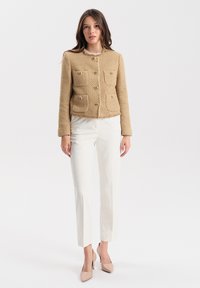 Beżowa kurtka z tweedu z czterema przednimi kieszeniami, okrągłym dekoltem i ozdobnymi zapięciami, zestawiona z białymi dopasowanymi spodniami i cielistymi szpilkami.