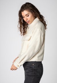 White fleece jacket with a collared design and front pocket, featuring long sleeves and a cropped fit, paired with dark jeans.
