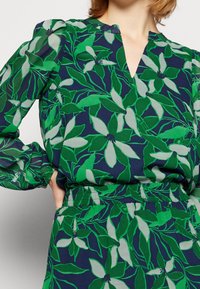 Groene en marineblauwe blouse met bloemenmotief, voorzien van een V-hals, pofmouwen en een gedetailleerde elastische tailleband. Lichtgewicht stof met een gladde textuur.