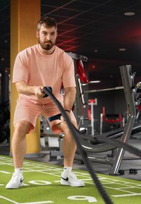 Homme athlétique en tenue rose clair, utilisant une corde de combat noire. Baskets blanches avec des accents noirs. Environnement de salle de sport avec gazon synthétique vert et équipements d'exercice.