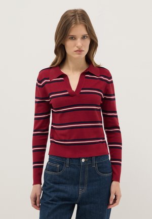 Vrouw met een rood shirt met lange mouwen met marineblauwe, witte en roze horizontale strepen en een blauwe spijkerbroek, staand tegen een effen witte achtergrond.