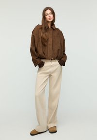 Soprabito in camoscio marrone con colletto e tasche ampie, abbinato a pantaloni crema a gamba larga e scarpe in camoscio marrone. Palette di colori semplice e neutra.