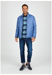 Helles blau gesteppte Jacke mit hohem Kragen, getragen über einem gestreiften Hemd. Eine dunkle Jeans und schwarze Sport Schuhe vervollständigen das Outfit.