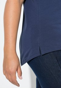 Zblízka pravá ruka uvolněná po boku, oblečená v námořnické modré košili a tmavě modrých džínách.