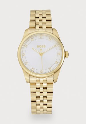 Montre en métal doré avec un cadran rond blanc, des marqueurs d'heure semblables à des diamants et des aiguilles dorées. Présente un bracelet à maillons dorés et un nom de marque gravé.