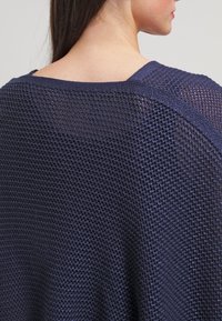 Mörkblå, löst stickad tröja med vid halsringning och strukturerad väv. Baksidan visar en avslappnad silhuett och mjukt, andningsbart tyg.