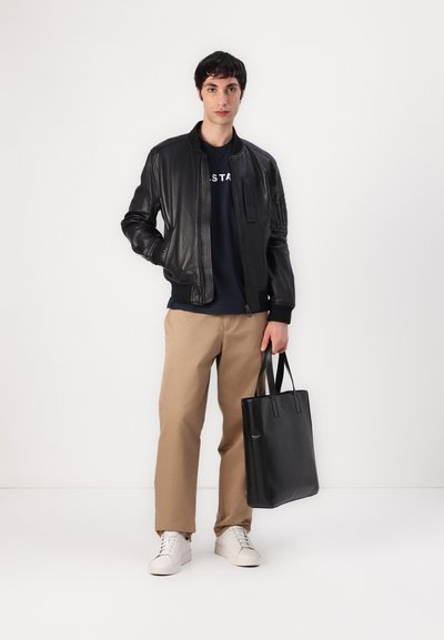 Veste bomber en cuir noir avec des poignets côtelés, t-shirt bleu marine avec des lettres, pantalon beige, baskets blanches, tenant un sac fourre-tout noir.