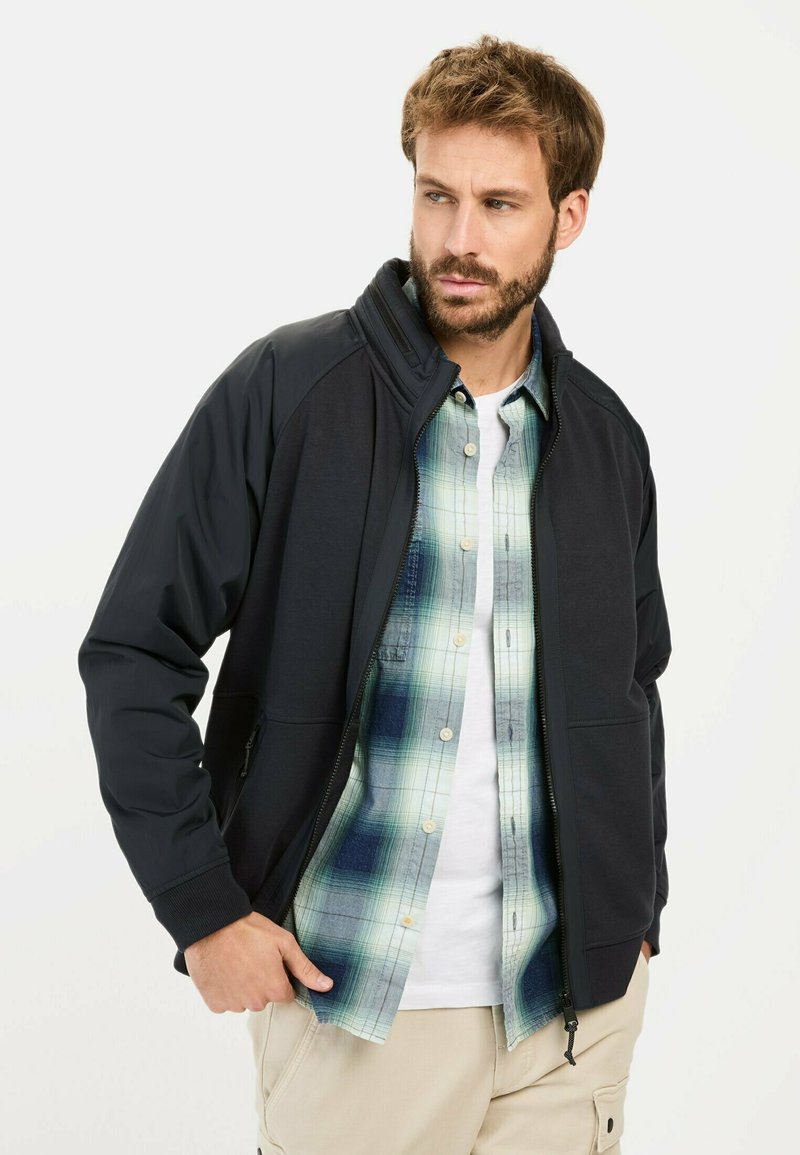Marine blauwe ritsjack met een staande kraag, gemaakt van zacht materiaal. Gedragen over een groen en blauwe geruite blouse, met beige cargobroeken.