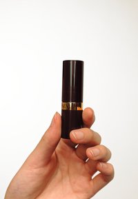 Ciemnofioletowa pomadka w tubce trzymana w dłoni, z złotą obrączką z napisem "Revlon". Gładka powierzchnia, cylindryczny kształt, błyszczące wykończenie.
