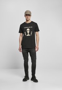 T-shirt en coton noir avec un graphique d'alien et le texte "JE VIENS EN PAIX", associé à un jean foncé et des baskets noires.