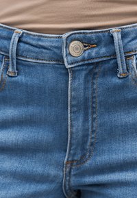 Μπλε τζιν παντελόνι στερεωμένο με μεταλλικό κουμπί, με εμφανείς ραφές και θηλιές για ζώνη, φορεμένο με μπεζ μπλούζα.