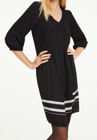 Schwarzes Kleid mit vertikalen Falten, V-Ausschnitt, 3/4-Ärmeln und einem ausgestellten Saum. Mit horizontalen weißen Streifen am Saum.