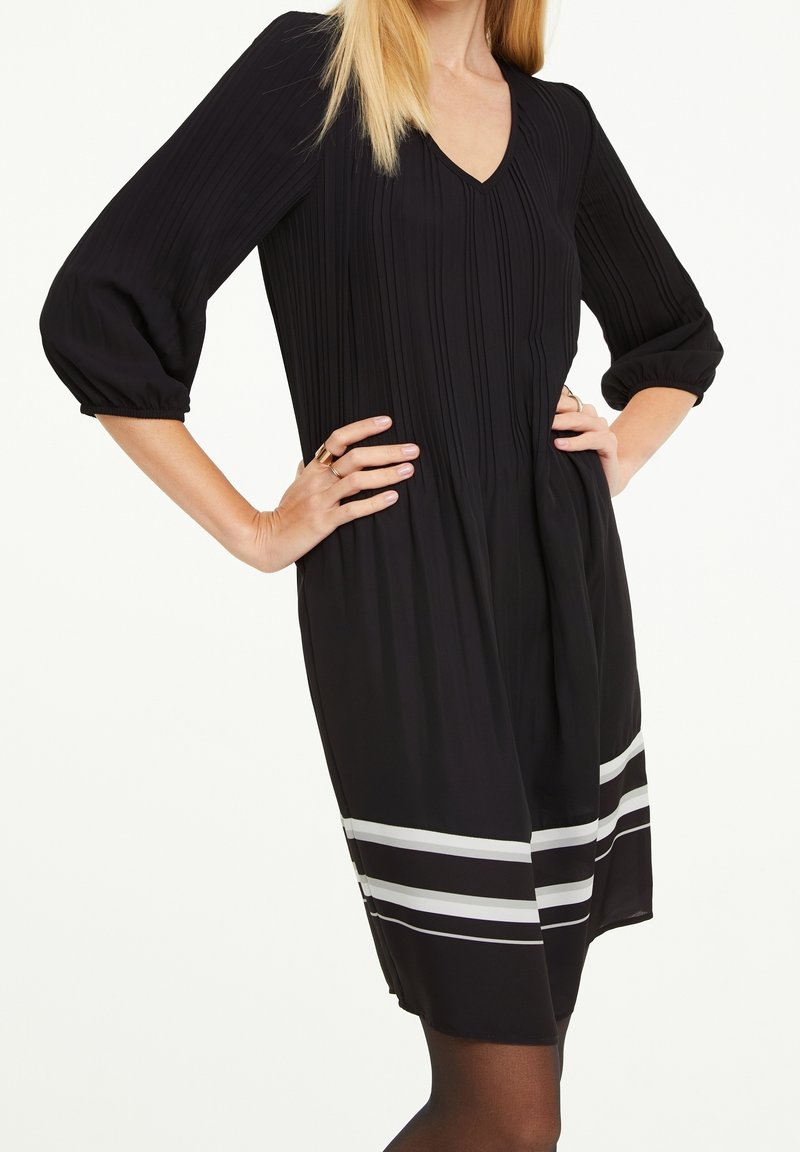 Schwarzes Kleid mit vertikalen Falten, V-Ausschnitt, 3/4-Ärmeln und einem ausgestellten Saum. Mit horizontalen weißen Streifen am Saum.
