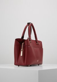 Rote Ledertasche mit strukturierter Oberfläche, zwei Griffen, einem goldfarbenen Reißverschlussdetail und einer strukturierten Form. Ausgestattet mit einer Vordertasche und dezentem Branding.