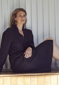 Schwarzer Bademantel mit Waffelstruktur, Taillengürtel, langen Ärmeln und Schalkragen. Über einem dunklen Unterkleid getragen, sitzt vor einem weißen Holzhintergrund.