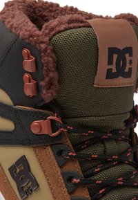 Încălțămintea DC verde și marron prezintă o suprafață texturată, un guler pufoșel, accente roșii și șireturi negre, având un design durabil pentru stabilitate.