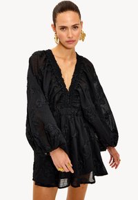 Vestido negro con escote en V profundo, ribete con volantes y mangas largas tipo globo. Presenta bordados florales y un dobladillo acampanado. Tejido ligero.