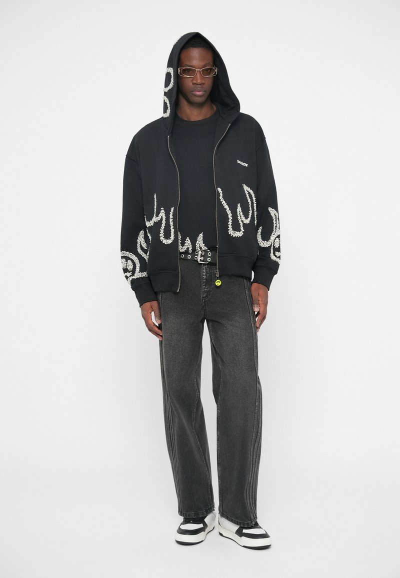 Άνδρας που φοράει μαύρη κουκούλα με λευκά σχέδια φλόγας, μαύρο μπλουζάκι, σκούρα γκρι τζιν, μαύρη ζώνη με μεταλλικές τρύπες, γυαλιά και μαύρα-λευκά αθλητικά παπούτσια.