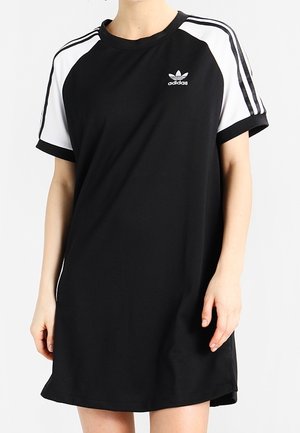 Vrouw draagt een zwart Adidas jurk met korte mouwen, witte panelen op de schouders en drie zwarte strepen op de mouwen.