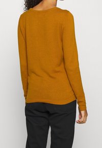 Pull en maille jaune moutarde avec un motif texturé, manches longues, col rond et ourlet côtelé. Porté avec un pantalon noir.
