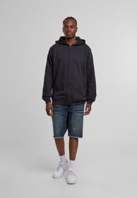 Tmavošedá mikina na zips s kapucňou so šnúrkou, nosená nad svetlomodrými džínsovými šortkami. Biele tenisky a ponožky po členky dotvárajú outfit.