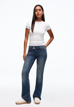 Nő áll, fehér, testhez simuló rövid ujjú inget, sötétkék, bő szárú farmert dupla gombos rögzítéssel, valamint fehér sportcipőt visel, fehér háttér előtt.