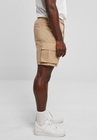 Shorts cargo beiges avec une coupe décontractée, dotés d'une poche latérale et d'une taille élastique. Associés à des baskets blanches et des chaussettes mi-mollet.