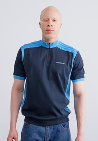 Melnā un zila polo krekls ar rāvējslēdzēju un ribotu tekstūru, ar trim zilām svītrām uz pleciem un Adidas logo uz krūts.