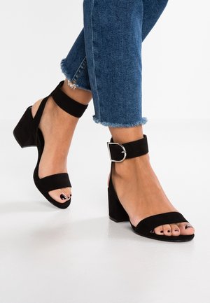Black suede block-heeled sandals with ankle straps. Features smooth straps and a square buckle. Worn with cropped blue jeans with frayed hems.
