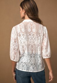 Balta mežģīņu blūze ar ziedu rakstiem, pūstām piedurknēm un vieglu, caurspīdīgu audumu. Dizains ietver smalkas detaļas uz aizmugures.