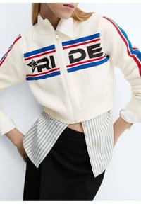 Krémový svetr na zip obsahuje výrazný text "RIDE" s modrými a červenými pruhy. Je překryt bílým a šedým pruhovaným tričkem.