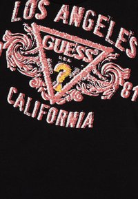 Svart bomull T-skjorte med slitt rød og hvit grafisk tekst med utskårne design, inkludert "Los Angeles" og "Guess" logoer.
