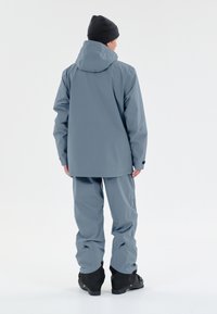 Lys grå vandtæt jakke med hætte og matchende bukser. Har justerbare manchetter og forstærkede sorte støvler. Glat tekstur, praktisk design.