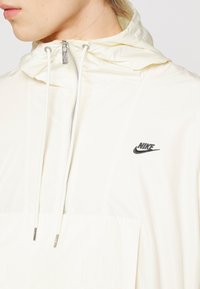 Nike pullover jakke i lyscreme med lynlåsning ved halsen, justerbar hætte og lille sort logo på brystet. Glat, let materiale.